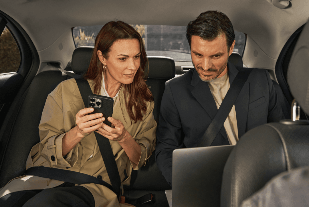 Two business travellers looking at phone with Freenow logo as red taxi pulls up